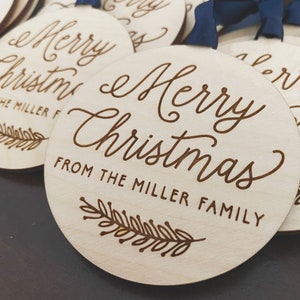 Puede incluir: Un conjunto de adornos redondos de madera con el texto "Merry Christmas" y "From the Miller Family" grabado en ellos. Cada adorno presenta un peque&ntilde;o dise&ntilde;o de pluma estilizado.