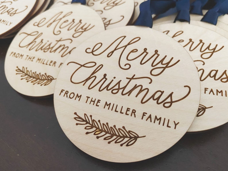Puede incluir: Un conjunto de adornos redondos de madera con el texto "Merry Christmas" y "From the Miller Family" grabado en ellos. Cada adorno presenta un peque&ntilde;o dise&ntilde;o de pluma estilizado.