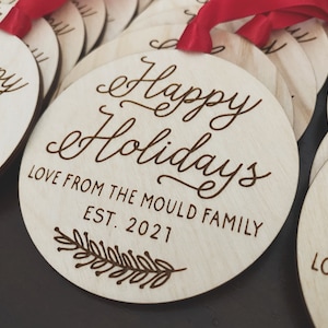 Puede incluir: Un conjunto de adornos redondos de madera con el texto "Happy Holidays" y "Love from the Mould Family Est. 2021" en una fuente cursiva. Cada adorno tiene una cinta roja.