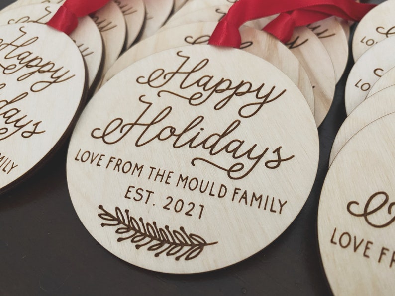 Puede incluir: Un conjunto de adornos redondos de madera con el texto "Happy Holidays" y "Love from the Mould Family Est. 2021" en una fuente cursiva. Cada adorno tiene una cinta roja.