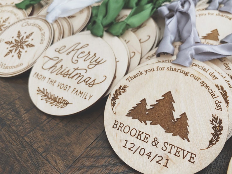 Op de afbeelding: Een verzameling ronde houten ornamenten met lasergegraveerde ontwerpen. Een ornament zegt "Merry Christmas from the Vogt Family". Een ander ornament heeft een ontwerp van drie dennenbomen en zegt "Brooke & Steve 12/04/21".