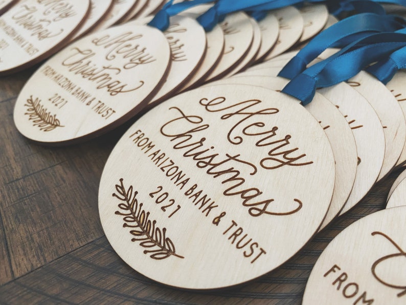 Puede incluir: Una pila de adornos redondos de madera con una cinta azul. Los adornos est&aacute;n grabados con el texto "Merry Christmas from Arizona Bank & Trust 2021".