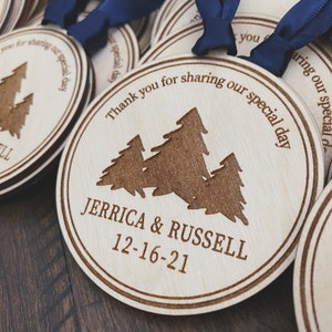 Op de afbeelding: Ronde houten ornamenten met een lasergegraveerd ontwerp van drie dennenbomen en de tekst "Bedankt voor het delen van onze speciale dag" en "Jerrica & Russell 12-16-21".