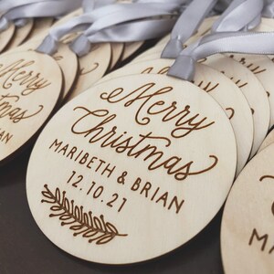 Puede incluir: Un conjunto de adornos navide&ntilde;os de madera con una cinta blanca. Los adornos est&aacute;n grabados con el texto "Merry Christmas" y los nombres "Maribeth & Brian" y la fecha "12.10.21".