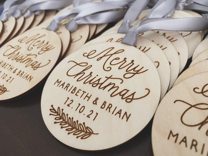 Puede incluir: Un conjunto de adornos navide&ntilde;os de madera con una cinta blanca. Los adornos est&aacute;n grabados con el texto "Merry Christmas" y los nombres "Maribeth & Brian" y la fecha "12.10.21".