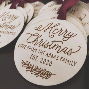 Puede incluir: Un conjunto de adornos redondos de madera con el texto "Merry Christmas" y "Love from the Abbas Family Est. 2020" grabado en ellos. Los adornos est&aacute;n decorados con una peque&ntilde;a rama de hojas.
