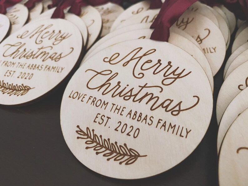 Puede incluir: Un conjunto de adornos redondos de madera con el texto "Merry Christmas" y "Love from the Abbas Family Est. 2020" grabado en ellos. Los adornos est&aacute;n decorados con una peque&ntilde;a rama de hojas.