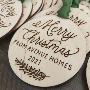 Puede incluir: Un conjunto de adornos redondos de madera con el texto "Merry Christmas from Avenue Homes 2021" grabado en ellos. Los adornos est&aacute;n decorados con una peque&ntilde;a rama de hojas.