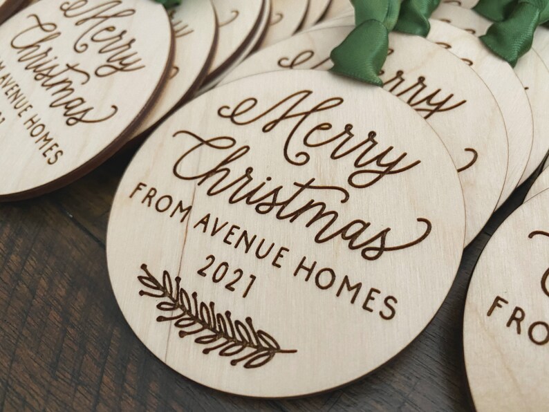 Puede incluir: Un conjunto de adornos redondos de madera con el texto "Merry Christmas from Avenue Homes 2021" grabado en ellos. Los adornos est&aacute;n decorados con una peque&ntilde;a rama de hojas.