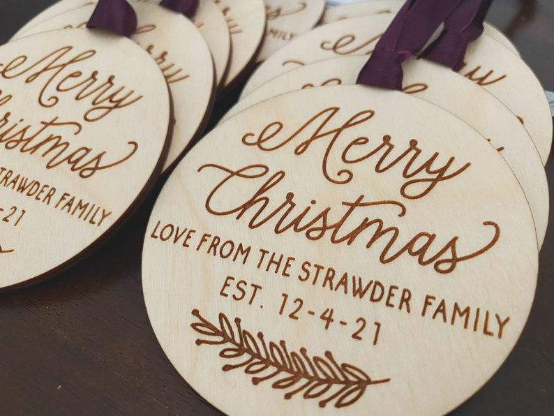 Puede incluir: Un conjunto de adornos redondos de madera con el texto "Merry Christmas" y "Love from the Strawder Family Est. 12-4-21" grabado en ellos. Los adornos son de color marr&oacute;n claro y tienen un aspecto r&uacute;stico y hecho a mano.