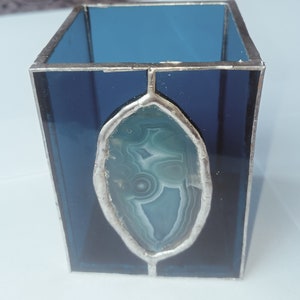 May include: A stained glass candle holder with a rectangular shape. The glass is a deep blue color, with a silver-colored metal frame. An oval-shaped, light blue and white agate stone is set in the center of the front panel.