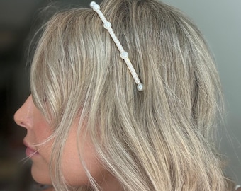May include: A pearl and crystal headband is worn in the hair. The headband features a thin, woven band with evenly spaced pearls and crystals. The hair is blonde and styled with soft waves. The background is blurred.