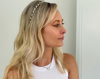 May include: A gold-toned headband with small, evenly spaced faux pearls. The headband is worn on blonde hair. The image shows a side profile of a person wearing a white tank top and a silver necklace with a square pendant.