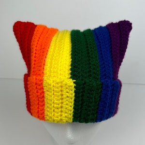 May include: A rainbow striped crochet beanie with cat ears. The beanie is made with red, orange, yellow, green, blue, and purple yarn.