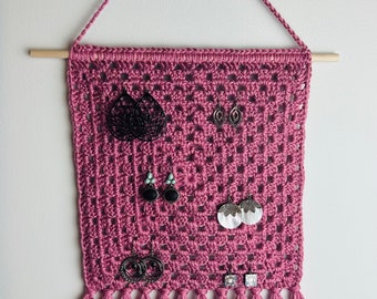 May include: A mauve crochet wall hanging organizer featuring a square grid pattern suspended from a wooden dowel with a matching yarn hanger. The textile display holds six pairs of various earrings, including black filigree, turquoise, and white shell designs, finished with a row of long fringe at the bottom.