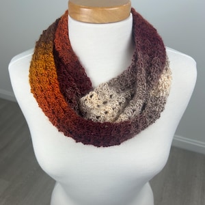 May include: A handmade infinity scarf in a gradient of warm colors, including orange, red, and beige. The crocheted scarf is displayed on a white mannequin, showcasing its textured stitch pattern and circular design. This accessory is perfect for adding a touch of handmade charm to any outfit.