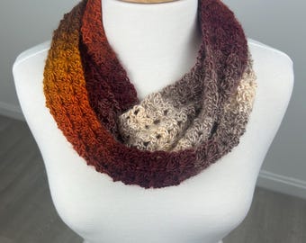 May include: A handmade infinity scarf in a gradient of warm colors, including orange, red, and beige. The crocheted scarf is displayed on a white mannequin, showcasing its textured stitch pattern and circular design. This accessory is perfect for adding a touch of handmade charm to any outfit.