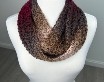 May include: A crocheted infinity scarf displayed on a mannequin. The scarf features a gradient of colors, transitioning from deep burgundy to shades of brown and tan. The open weave design is visible, showcasing the texture of the yarn.