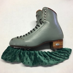 Puede incluir: Un par de patines de hielo grises con un tacón marrón y un protector de patines de terciopelo verde.