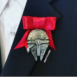 Könnte beinhalten: Eine rote Fliege mit einer silbernen Millennium Falcon-Anstecknadel. Die Anstecknadel ist eine detaillierte Nachbildung des legendären Raumschiffs aus Star Wars.