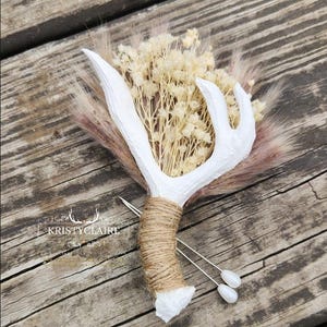 May include: A boutonniere featuring a white antler piece wrapped in tan twine, accented with dried cream-colored baby's breath and soft brown pampas grass. Two silver pins with white pearl-like heads are attached. The text KRISTYCLAIRE is visible.