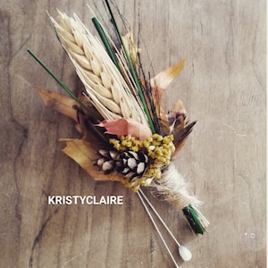 May include: A rustic boutonniere featuring wheat, dried leaves, a pine cone, and small yellow flowers, wrapped with twine. Two white-tipped stems extend from the base. The text "KRISTYCLAIRE" is visible on the wooden background.