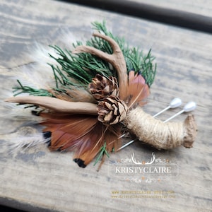 May include: A rustic wedding boutonniere featuring a brown antler, pine cones, brown feathers, and green foliage. The boutonniere is secured with twine and two silver pins.