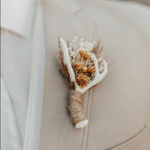 Op de afbeelding: Een witte geweiboutonnière met gedroogde bloemen en een bruine touw strik. De boutonnière is vastgemaakt aan een beige colbert.