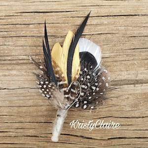 May include: A small bouquet of feathers in black, white, and gold with a silver pin. The feathers are arranged in a circular shape and have a spotted pattern.