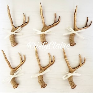 May include: Six gold deer antlers with white ribbon bows tied around the base of each antler. The antlers are wrapped in twine.