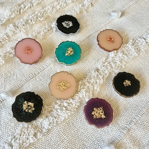 Könnte beinhalten: Sortiment von acht runden Handygriffen in verschiedenen Farben mit goldenen Glitzerakzenten. Die Handygriffe sind aus Harz gefertigt und haben eine glatte, glänzende Oberfläche.