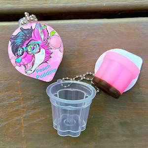 May include: A clear plastic dessert cup and two keychains. One keychain is a pink oval with a cartoon furry character and the words "Peach Pudding". The other keychain is a pink and brown dessert cup.
