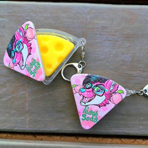 May include: A novelty keychain featuring a pink, triangular design with a cartoon fox character wearing glasses and the text "Peach Soda." The keychain opens to reveal a yellow, cheese-shaped insert. The design includes a silver-colored keyring.
