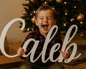 May include: A large, white wooden sign spelling out the name "Caleb" in a cursive font. The sign is held by a child, who is laughing. A Christmas tree with golden ornaments and lights is in the background.