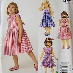 May include: A McCall's sewing pattern for girls' dresses. The pattern features illustrations of three different dress styles, including a red and white gingham dress, a blue plaid dress, and a floral print dress. The pattern includes the text "EASY stitch 'n save".