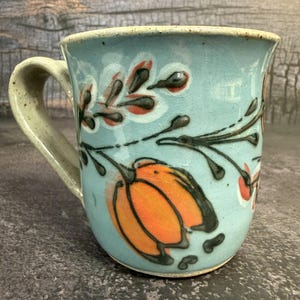 May include: A light blue ceramic mug with a speckled finish and a curved handle. The exterior features a hand-painted design of orange tulip-like flowers and dark brown leafy branches. The glaze has a smooth texture with visible dark speckles.