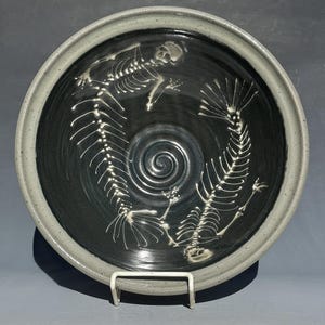 May include: A ceramic bowl with a black interior and a gray rim. The bowl features a white design of two fish skeletons. The skeletons are arranged in a circular pattern around a spiral in the center of the bowl.
