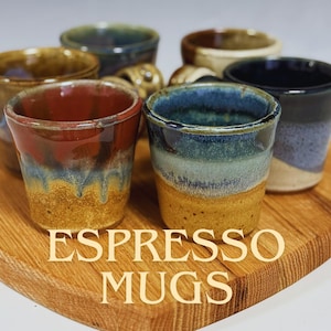 May include: A collection of handmade espresso mugs in various colors, including red, blue, and brown, displayed on a wooden serving board. The mugs have a rustic, glazed finish. The text "ESPRESSO MUGS" is printed on the board.