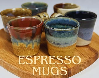 May include: A collection of handmade espresso mugs in various colors, including red, blue, and brown, displayed on a wooden serving board. The mugs have a rustic, glazed finish. The text "ESPRESSO MUGS" is printed on the board.