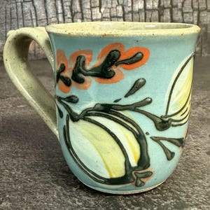 May include: A ceramic mug with a light blue glaze featuring a hand-painted design of dark green stems, yellow bulbous shapes, and orange leaf-like accents. The mug has a sturdy handle and a speckled texture throughout the light blue surface.