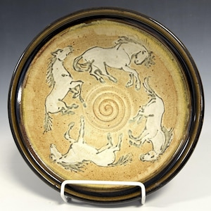 May include: A round ceramic plate with a tan, speckled surface featuring a central spiral design and three white horse figures arranged in a circular pattern. The plate has a dark brown rim and a smooth, glossy finish throughout the piece.