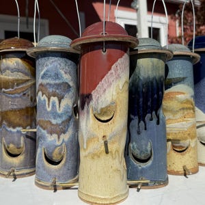 May include: A collection of handmade ceramic bird feeders in various colors, including blue, brown, and red. Each feeder has a unique design and a metal hanging wire. The feeders are cylindrical with a curved opening for birds to access food.