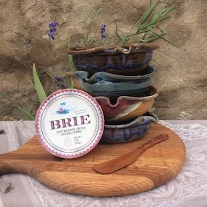 May include: A round, pink and white brie cheese package with the text "Happy Taste" and "BRIE" in a white box. The package is sitting on a wooden cutting board with a wooden butter knife. There are four stacked ceramic bowls with brown, blue, and pink glaze.