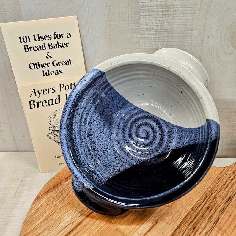 Pu&ograve; includere: Una ciotola per pane in ceramica con smalto bicolore, interno bianco sporco maculato ed esterno blu profondo. La ciotola presenta un motivo a spirale alla base ed &egrave; posizionata accanto a un libro intitolato 101 Uses for a Bread Baker & Other Great Ideas.