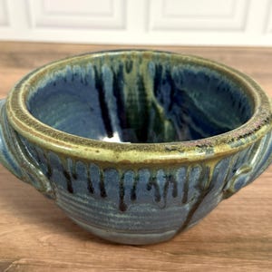 Pu&ograve; includere: Una ciotola in ceramica con due manici laterali, caratterizzata da uno smalto interno ed esterno blu intenso con gocciolature blu scuro e verdi lungo i lati. Il bordo &egrave; rifinito con uno smalto testurizzato color verde oliva.