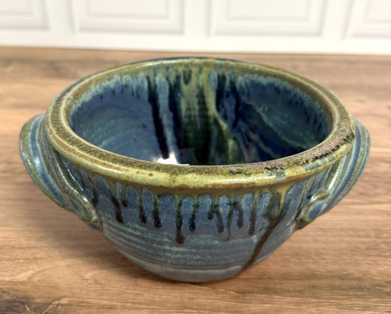 Pu&ograve; includere: Una ciotola in ceramica con due manici laterali, caratterizzata da uno smalto interno ed esterno blu intenso con gocciolature blu scuro e verdi lungo i lati. Il bordo &egrave; rifinito con uno smalto testurizzato color verde oliva.