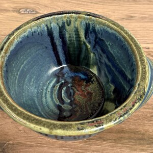Pu&ograve; includere: Una ciotola in ceramica rotonda con due piccole maniglie laterali, caratterizzata da uno smalto blu profondo e verde scuro con accenti marroni. La superficie interna presenta una finitura lucida con gocciolature.