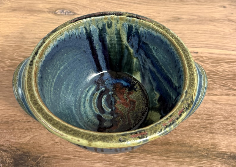 Pu&ograve; includere: Una ciotola in ceramica rotonda con due piccole maniglie laterali, caratterizzata da uno smalto blu profondo e verde scuro con accenti marroni. La superficie interna presenta una finitura lucida con gocciolature.