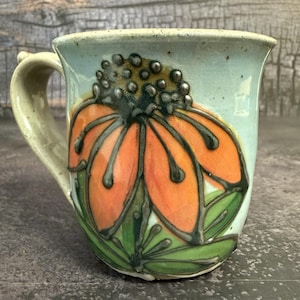 May include: A ceramic mug with a light blue speckled glaze featuring a large, hand-painted orange flower with dark green outlines and a textured, dotted center. The mug has a curved handle and a flared rim, showcasing a glossy finish throughout.