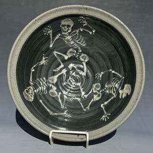 May include: A ceramic bowl with a dark gray interior and a light gray rim. The bowl features a white skeletal design of dancing skeletons. The bowl is displayed on a white metal stand. A Halloween-themed decorative item.